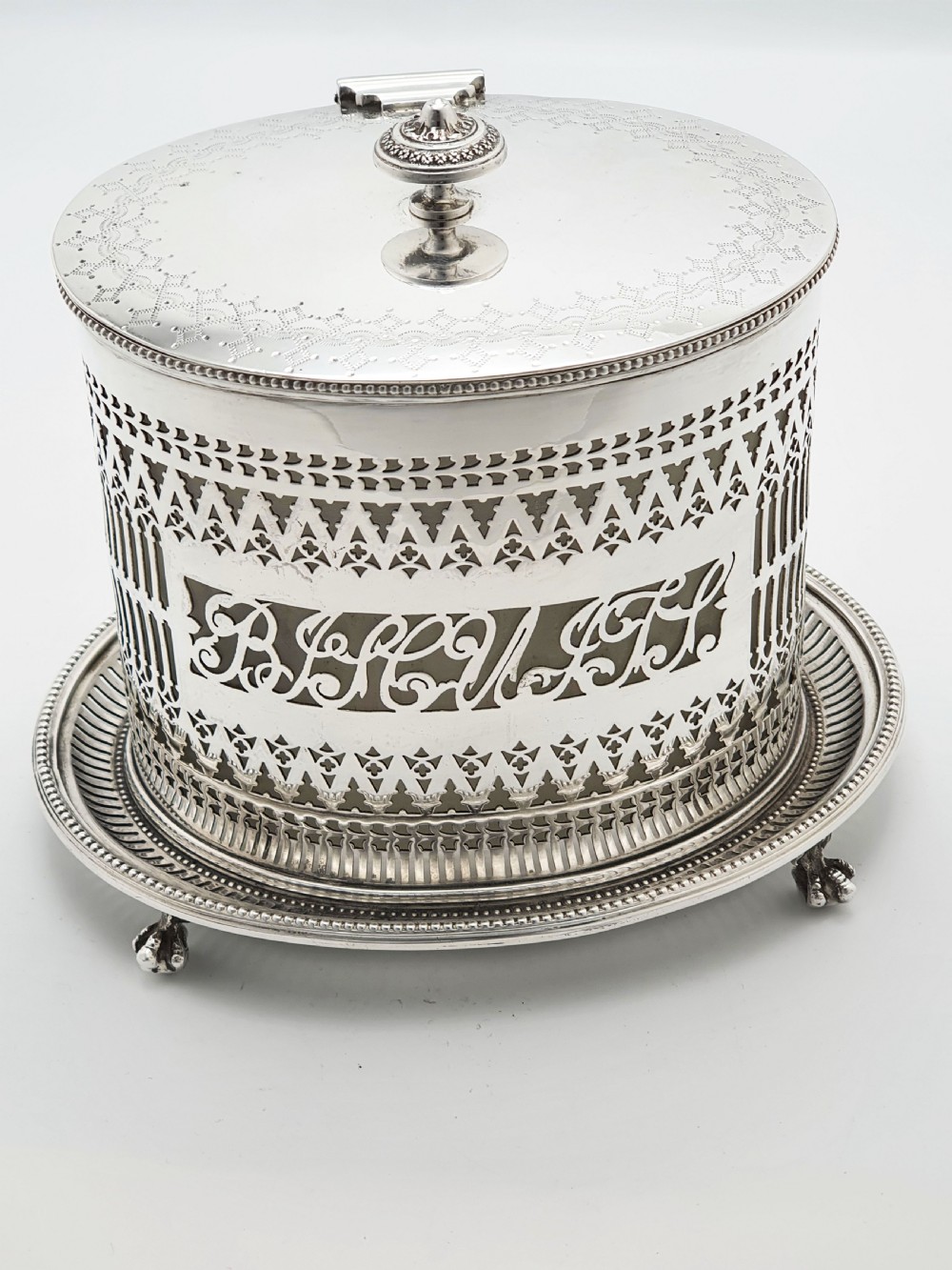 An Edwardian silver plated biscuit barrel featuring pierced scrollwork spelling out the word biscuits, a matching tray base, and a hinged lid with a decorative finial. View V