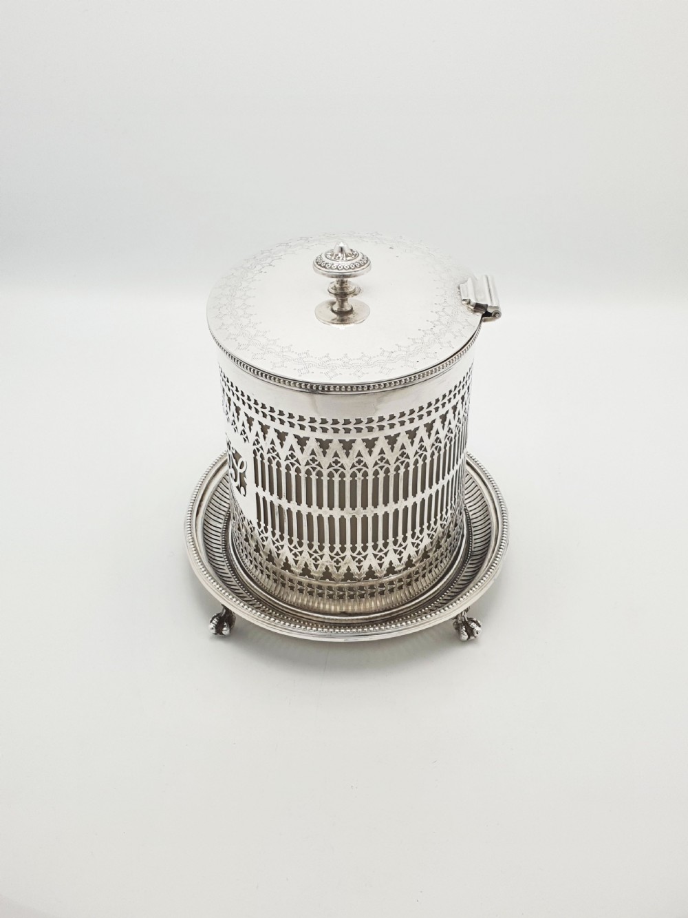 An Edwardian silver plated biscuit barrel featuring pierced scrollwork spelling out the word biscuits, a matching tray base, and a hinged lid with a decorative finial. View VI