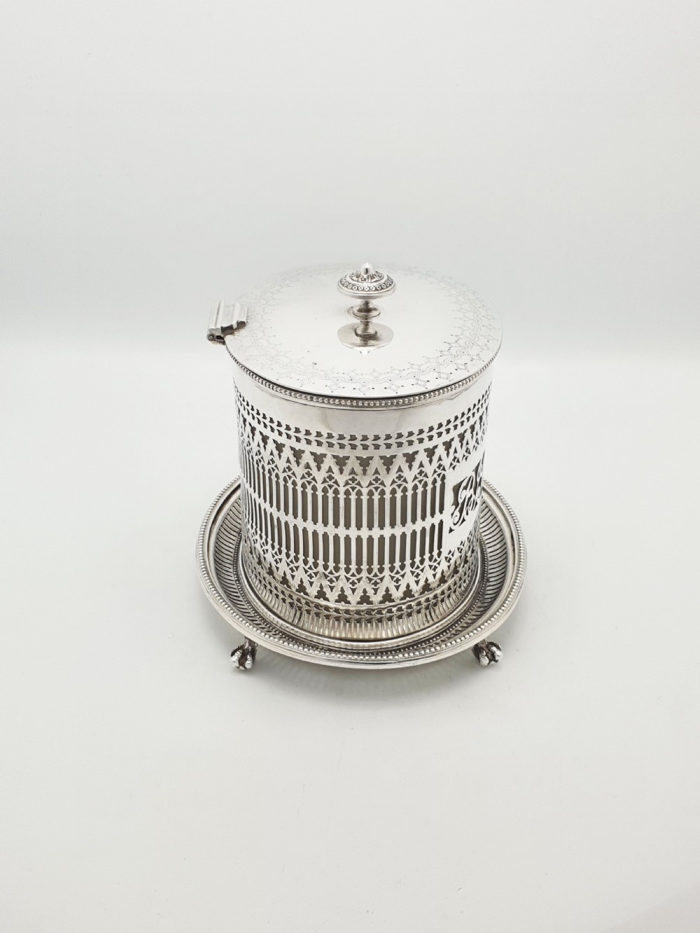 An Edwardian silver plated biscuit barrel featuring pierced scrollwork spelling out the word biscuits, a matching tray base, and a hinged lid with a decorative finial. View VIII