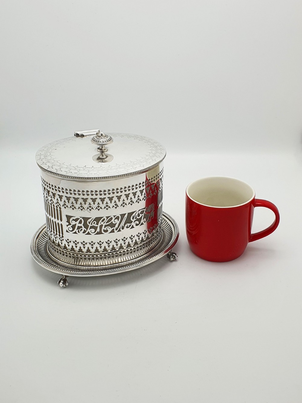 An Edwardian silver plated biscuit barrel featuring pierced scrollwork spelling out the word biscuits, a matching tray base, and a hinged lid with a decorative finial. View IX
