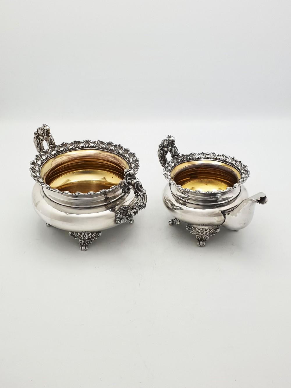A Georgian silver-plated sugar bowl and matching cream jug with heavily carved scroll rims, double-scroll handles, and gilded interiors.