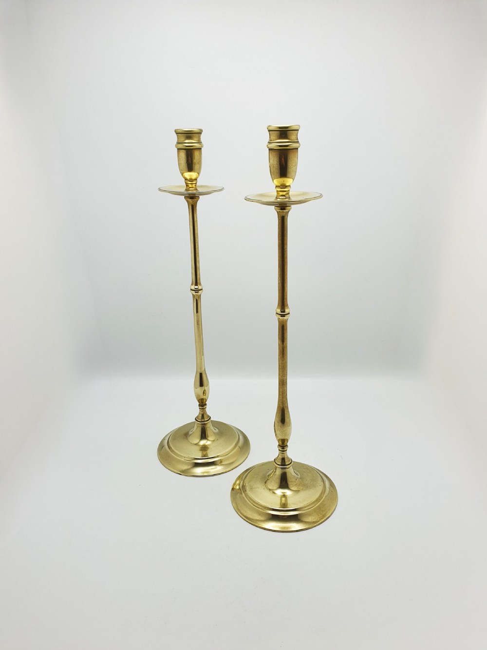 A pair of tall antique Edwardian brass candlesticks featuring slender turned stems, wide drip pans, and circular stepped bases.