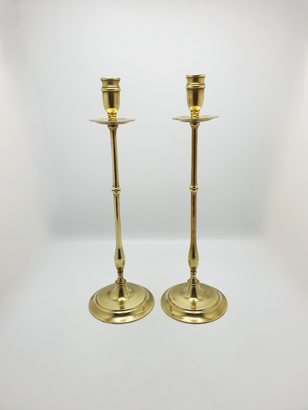A pair of tall antique Edwardian brass candlesticks featuring slender turned stems, wide drip pans, and circular stepped bases. View II