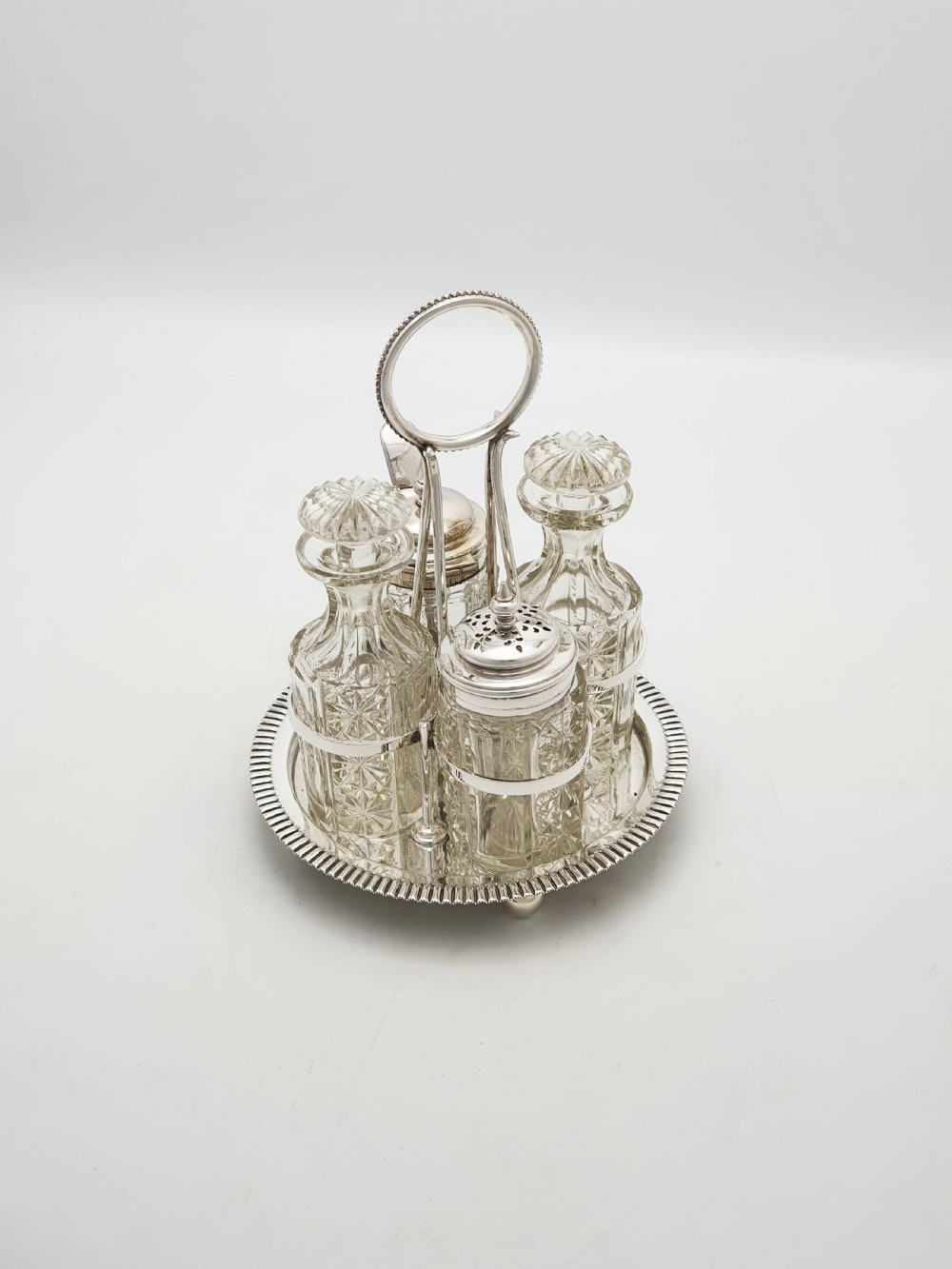 An antique silver-plated bachelor cruet set featuring cut-glass bottles on a circular gadrooned tray with a central carrying handle.