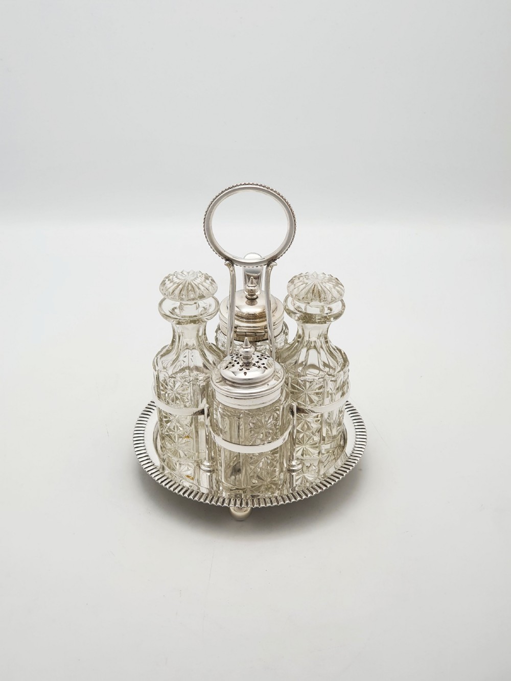 An antique silver-plated bachelor cruet set featuring cut-glass bottles on a circular gadrooned tray with a central carrying handle. View II