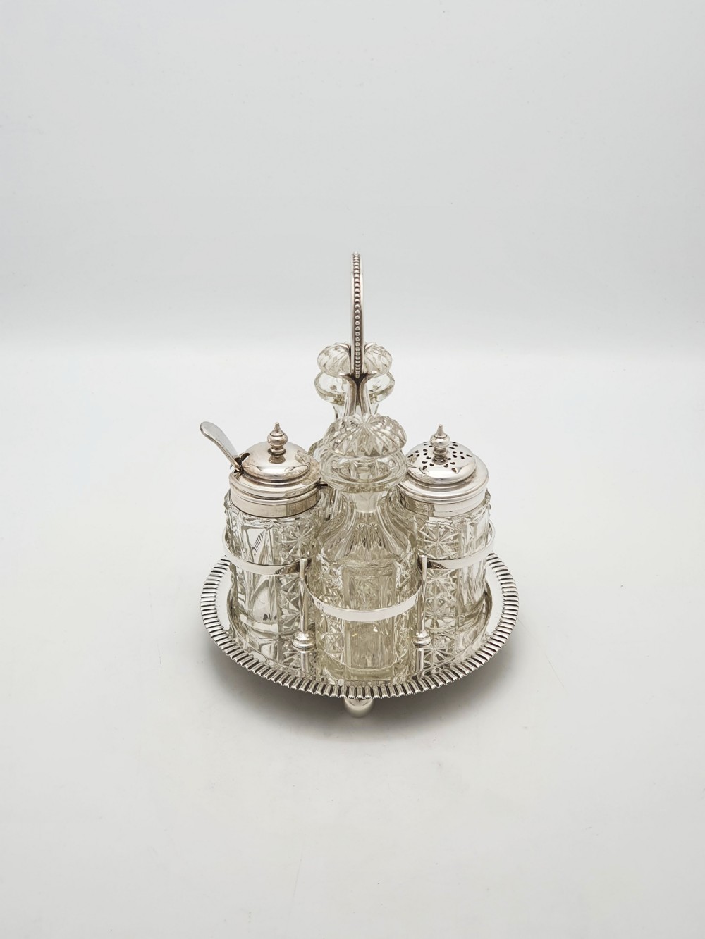 An antique silver-plated bachelor cruet set featuring cut-glass bottles on a circular gadrooned tray with a central carrying handle. View V