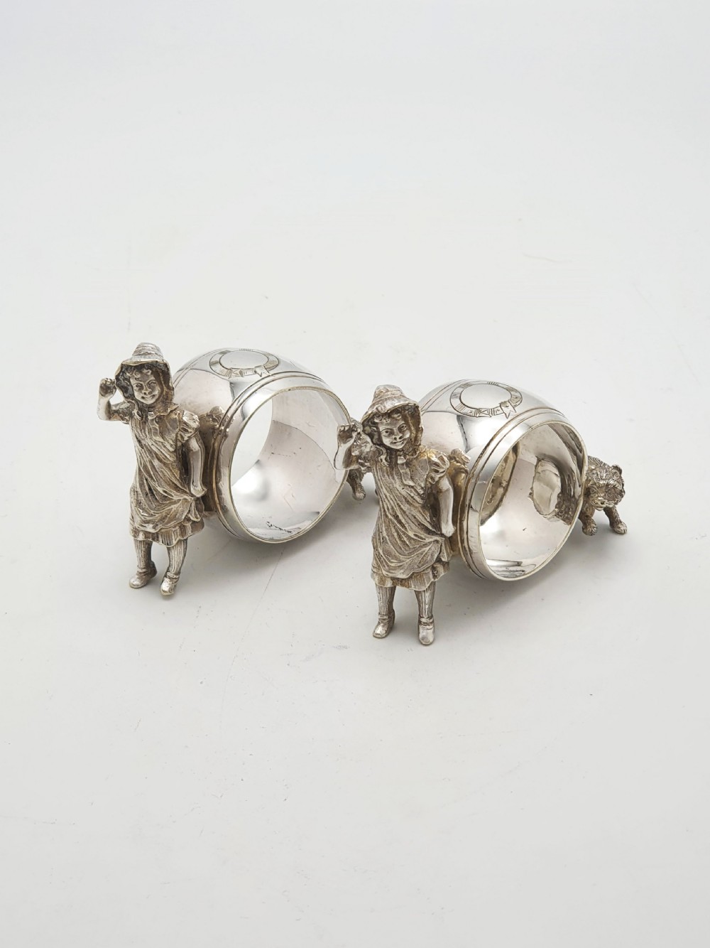 A pair of Victorian silver-plated napkin rings featuring detailed cast figures of young girls wearing bonnets standing beside engraved circular rings.