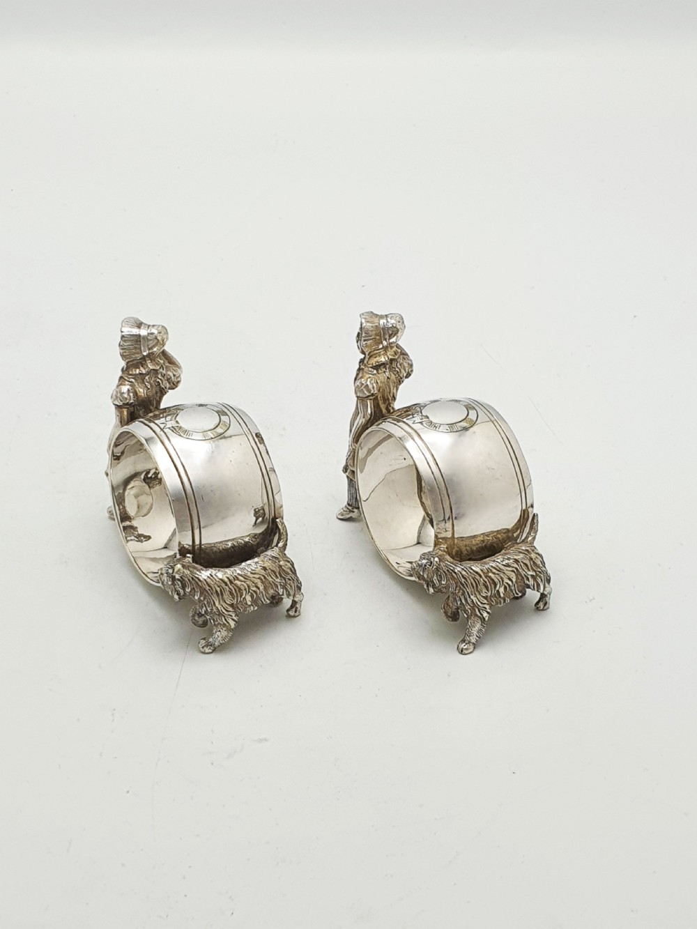 A pair of Victorian silver-plated napkin rings featuring detailed cast figures of young girls wearing bonnets standing beside engraved circular rings. View III