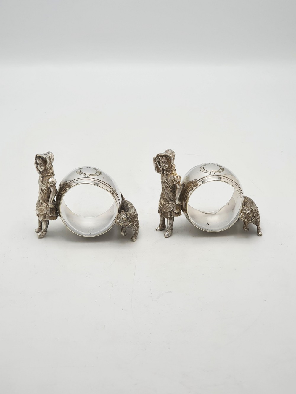 A pair of Victorian silver-plated napkin rings featuring detailed cast figures of young girls wearing bonnets standing beside engraved circular rings. View IV