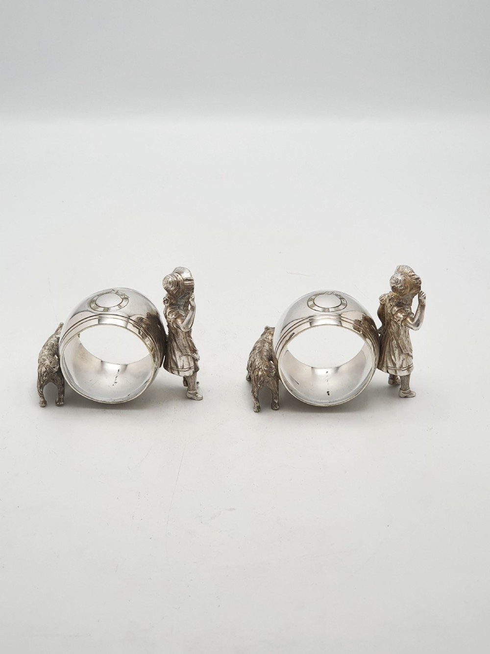 A pair of Victorian silver-plated napkin rings featuring detailed cast figures of young girls wearing bonnets standing beside engraved circular rings. View V
