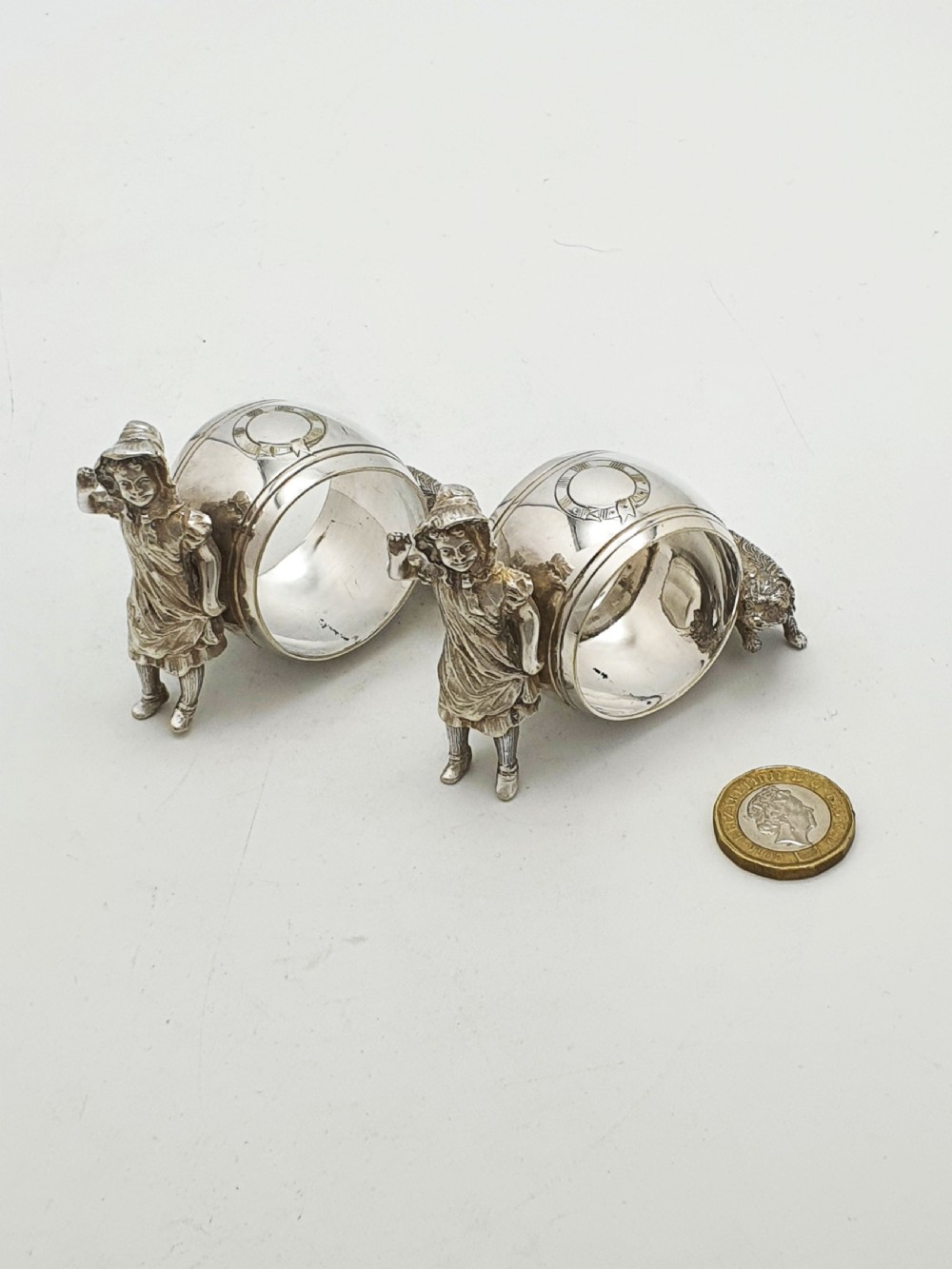 A pair of Victorian silver-plated napkin rings featuring detailed cast figures of young girls wearing bonnets standing beside engraved circular rings. View VII
