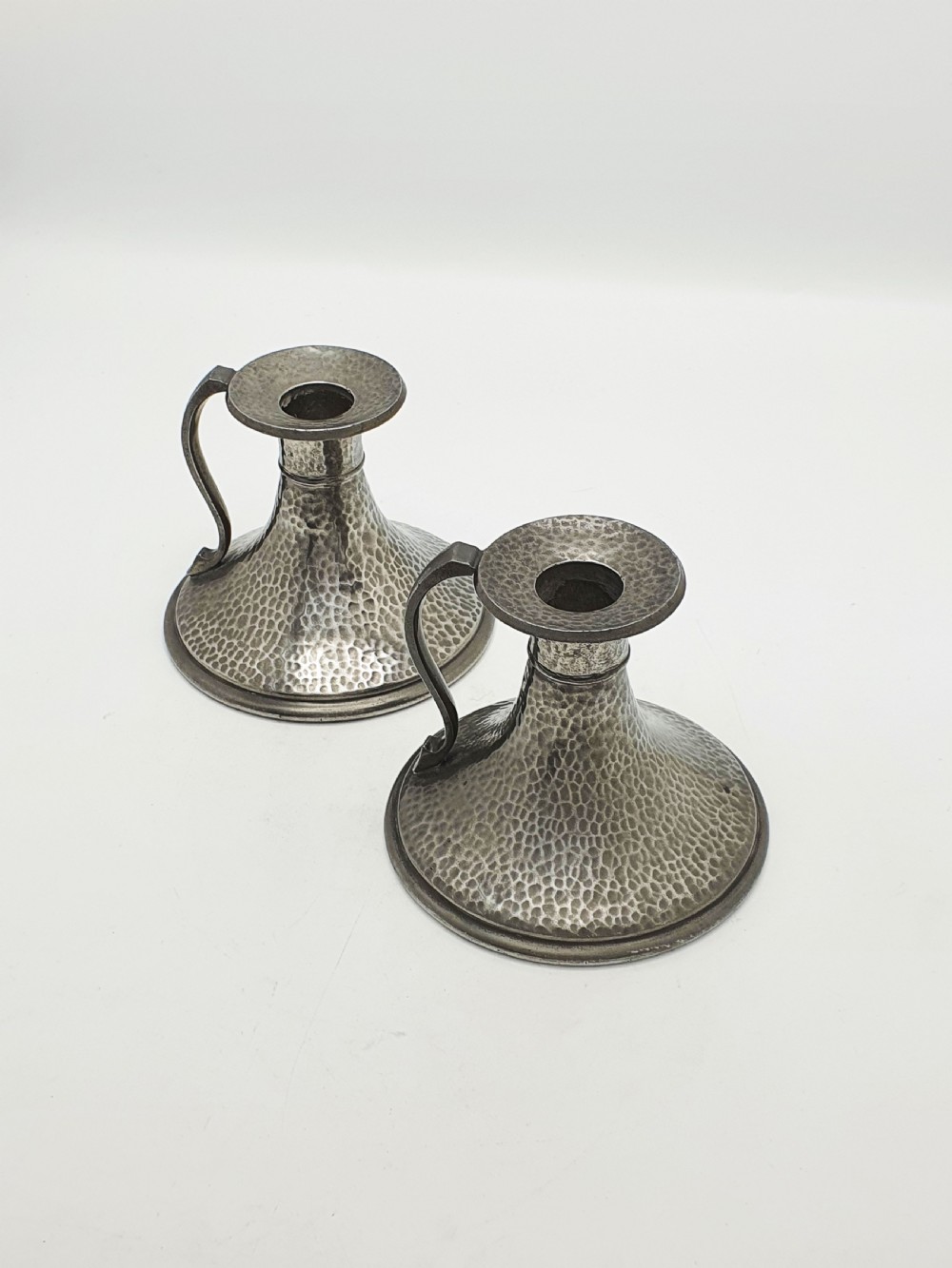 A pair of Arts and Crafts era flared pewter chambersticks with hand-hammered textures and curved side handles.