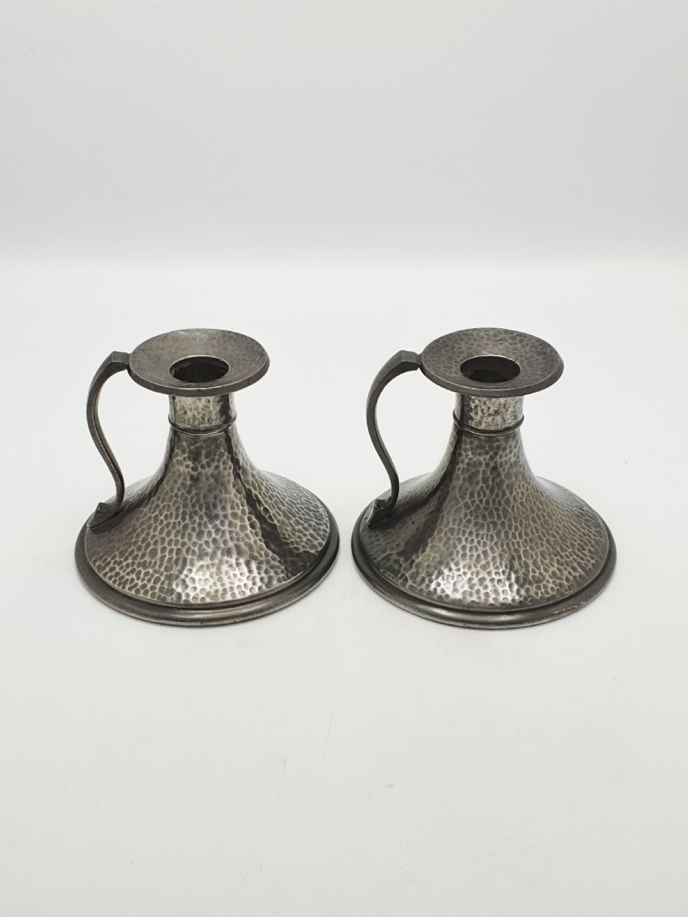 A pair of Arts and Crafts era flared pewter chambersticks with hand-hammered textures and curved side handles. View II