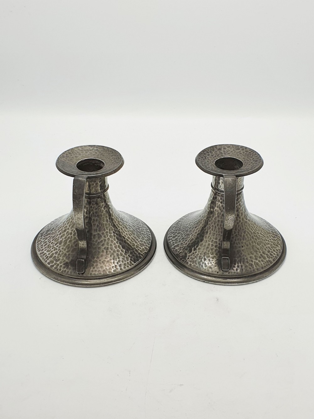 A pair of Arts and Crafts era flared pewter chambersticks with hand-hammered textures and curved side handles. View V