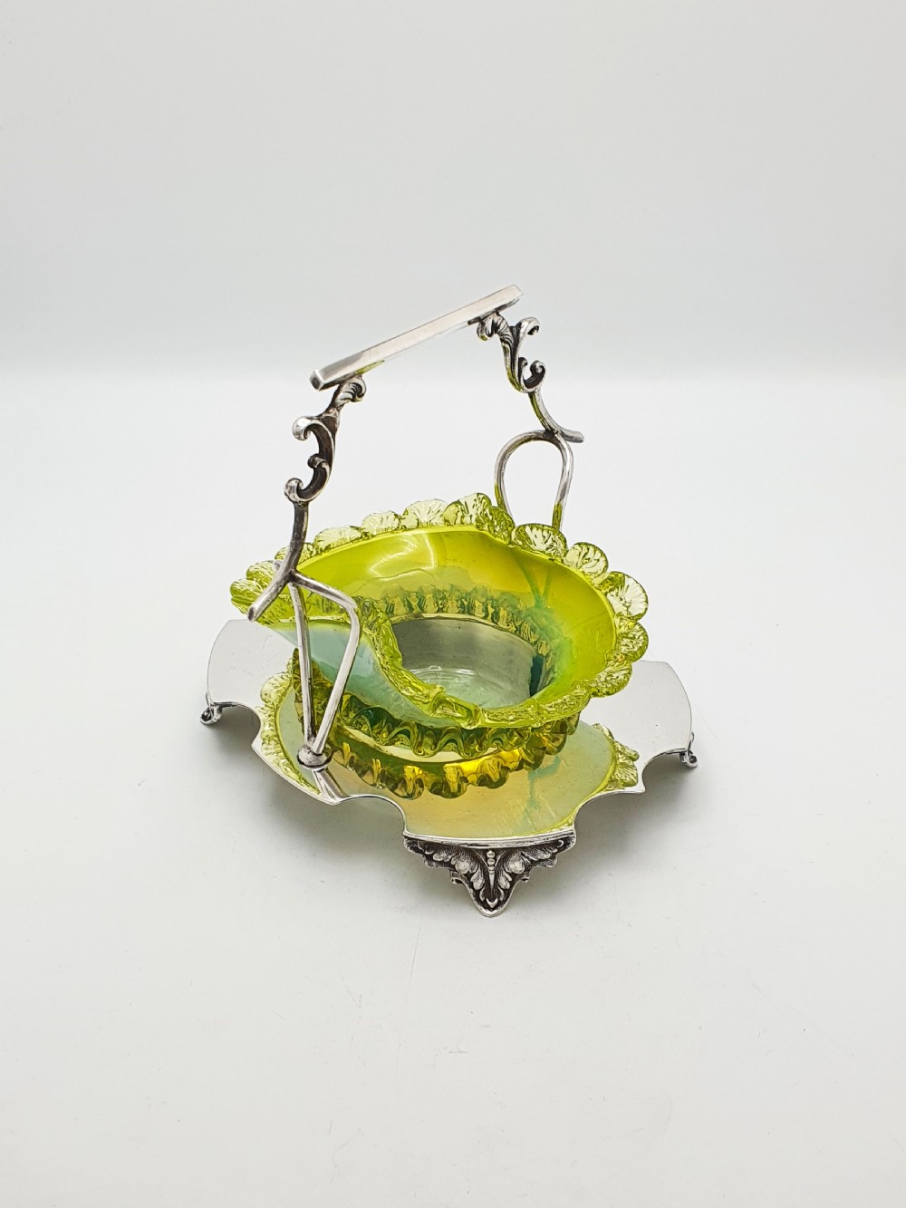 An Edwardian yellow-green vaseline glass bowl with a frilled rim set within a silver-plated frame featuring an overhead handle and ornamental feet.