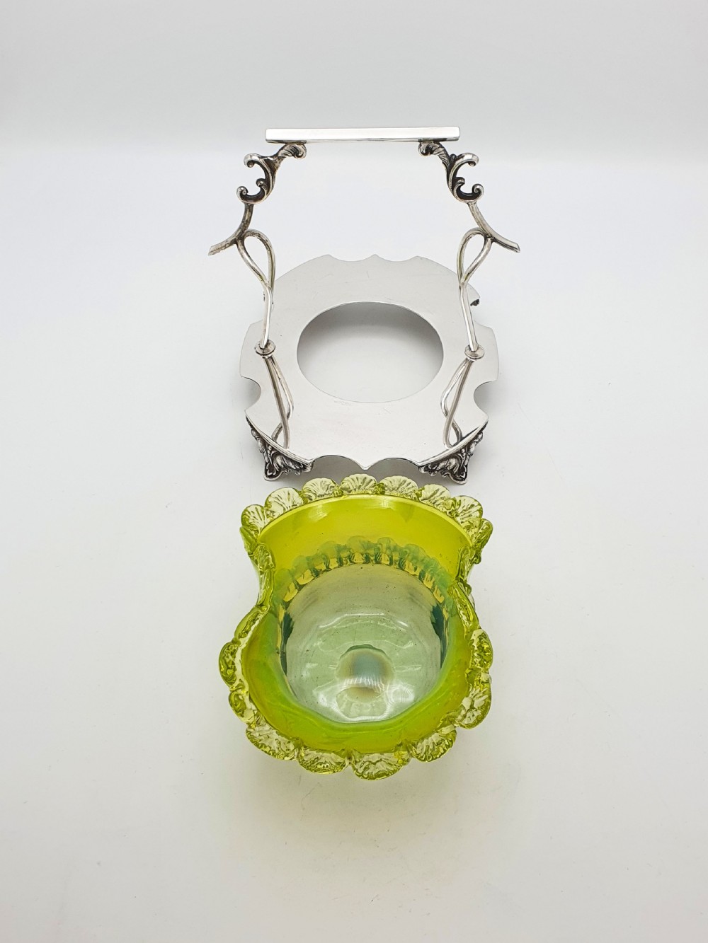 An Edwardian yellow-green vaseline glass bowl with a frilled rim set within a silver-plated frame featuring an overhead handle and ornamental feet. View II
