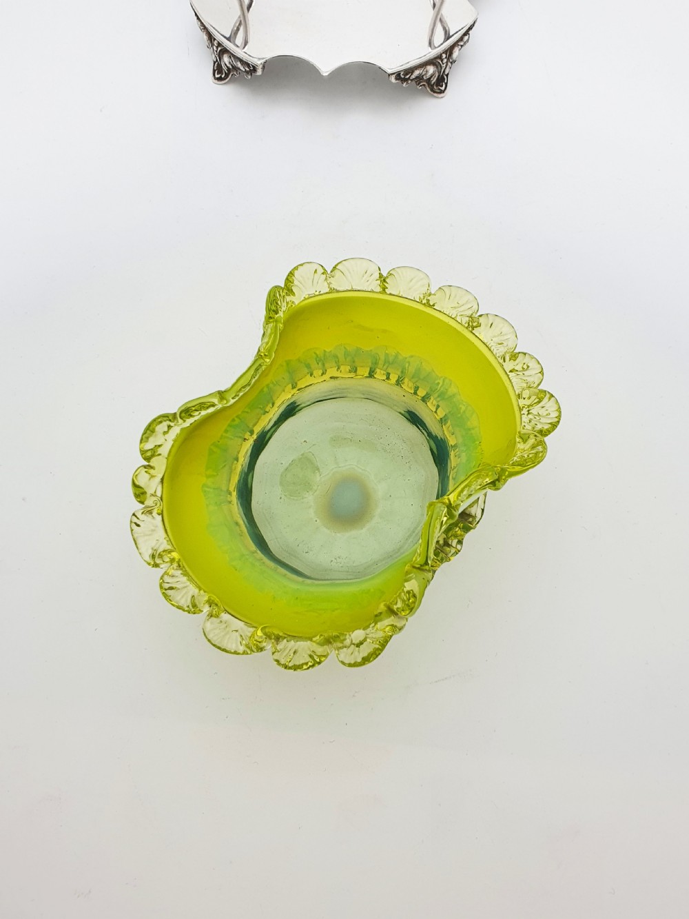 An Edwardian yellow-green vaseline glass bowl with a frilled rim set within a silver-plated frame featuring an overhead handle and ornamental feet. View III