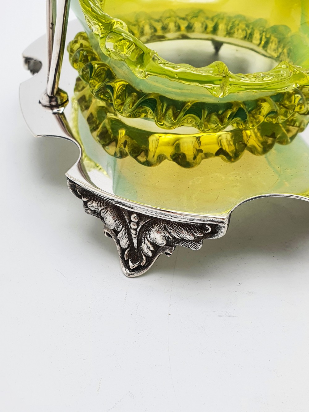 An Edwardian yellow-green vaseline glass bowl with a frilled rim set within a silver-plated frame featuring an overhead handle and ornamental feet. View VII