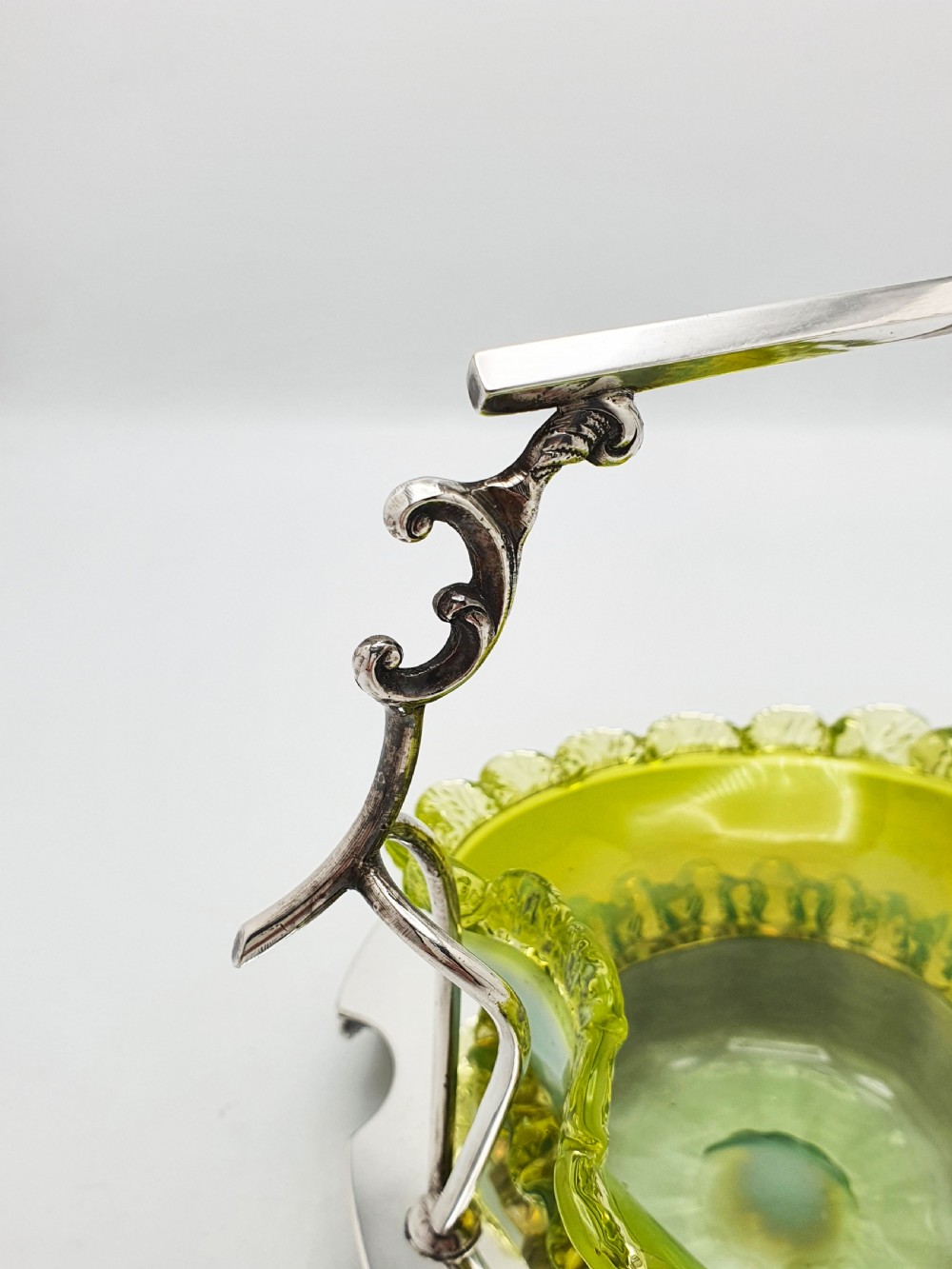 An Edwardian yellow-green vaseline glass bowl with a frilled rim set within a silver-plated frame featuring an overhead handle and ornamental feet. View VIII