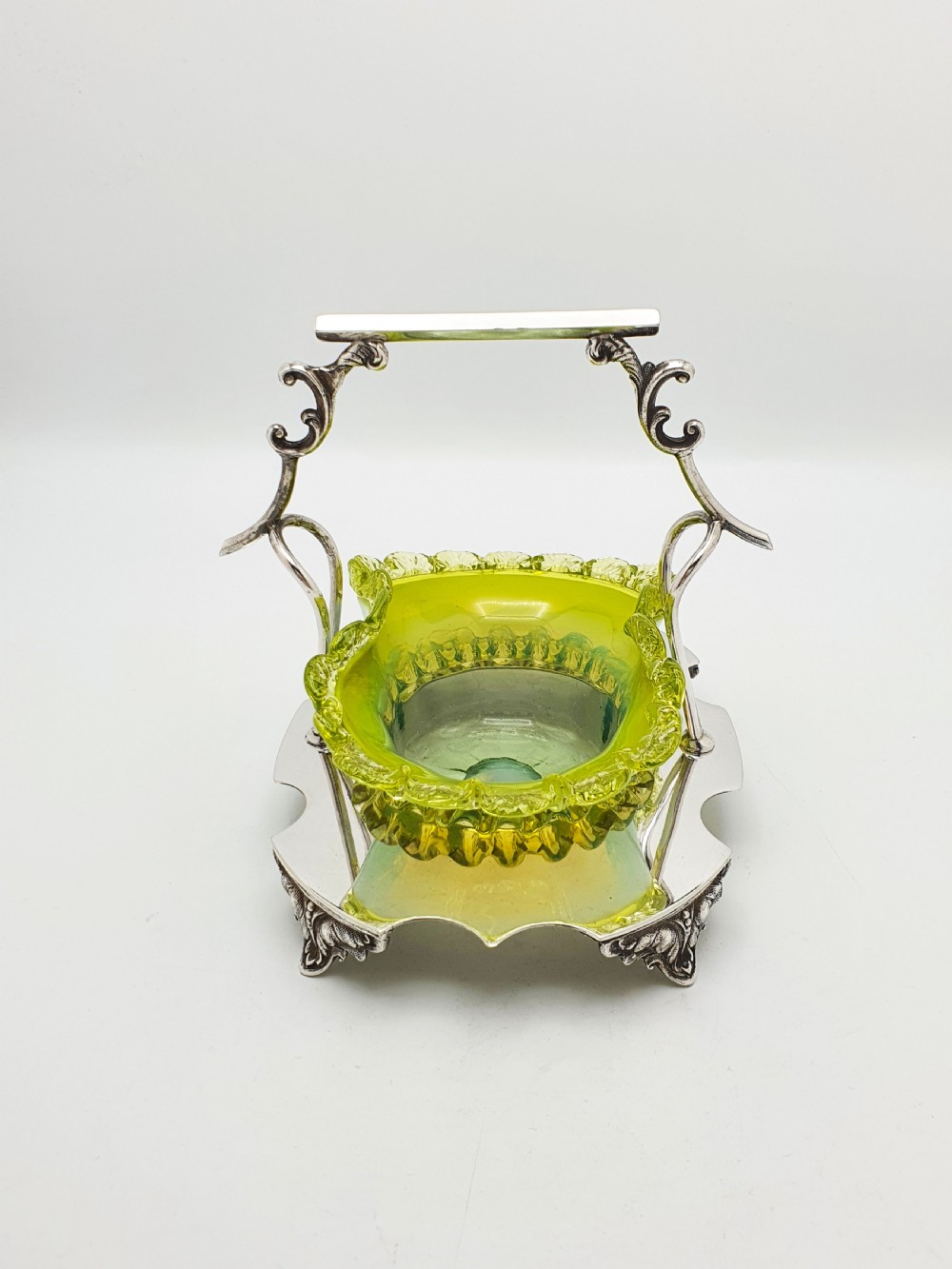 An Edwardian yellow-green vaseline glass bowl with a frilled rim set within a silver-plated frame featuring an overhead handle and ornamental feet. View IX