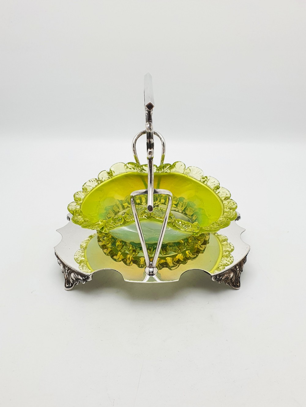 An Edwardian yellow-green vaseline glass bowl with a frilled rim set within a silver-plated frame featuring an overhead handle and ornamental feet. View X