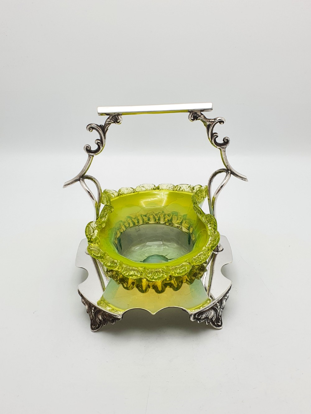 An Edwardian yellow-green vaseline glass bowl with a frilled rim set within a silver-plated frame featuring an overhead handle and ornamental feet. View XI