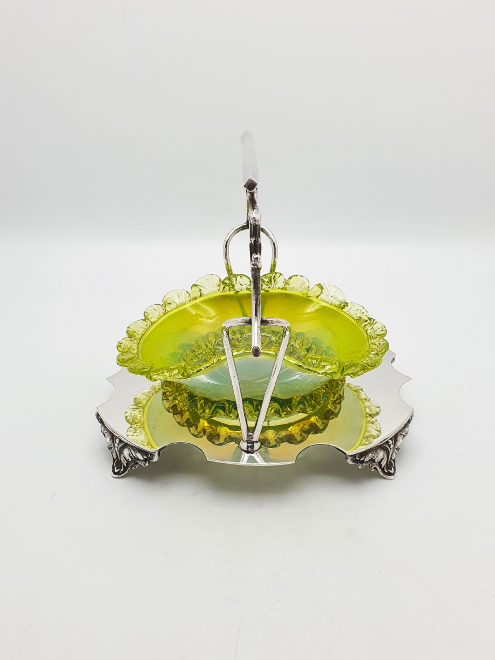 An Edwardian yellow-green vaseline glass bowl with a frilled rim set within a silver-plated frame featuring an overhead handle and ornamental feet. View XII
