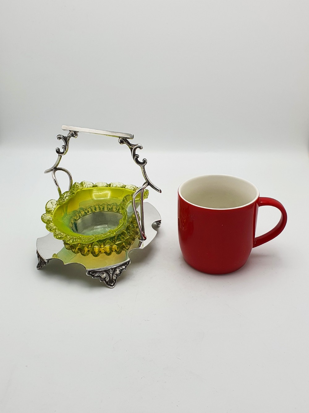 An Edwardian yellow-green vaseline glass bowl with a frilled rim set within a silver-plated frame featuring an overhead handle and ornamental feet. View XIII
