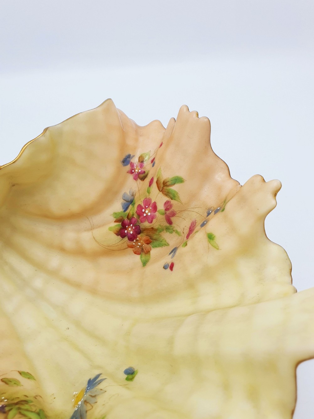A shell-shaped antique blush ivory porcelain dish featuring soft peach shading, hand-painted pink floral sprays, textured ridges, and a gilded scalloped edge. View III