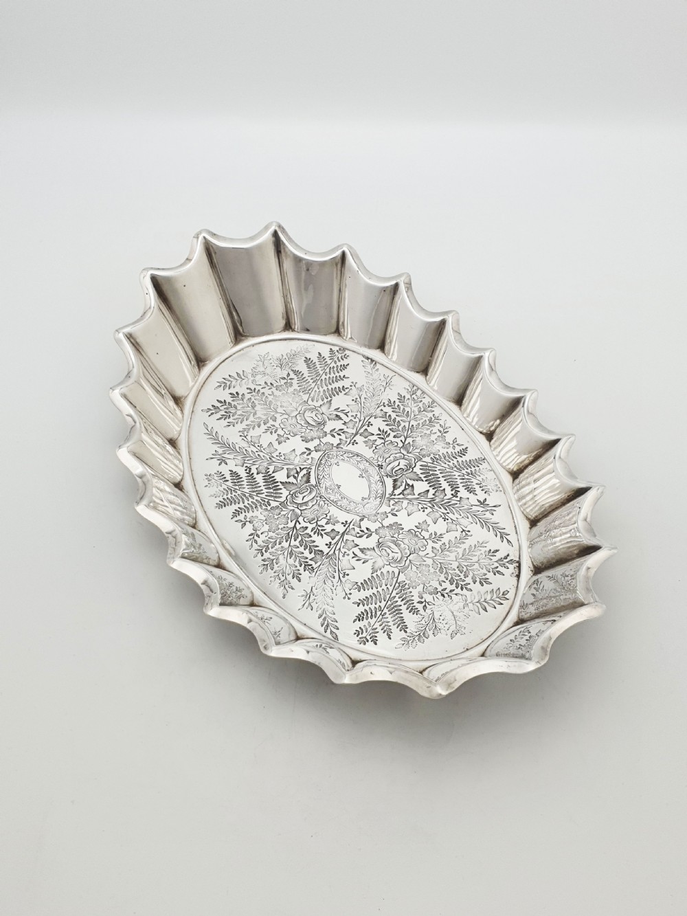 An oval silver plate serving dish featuring intricate floral engravings in the centre and a fluted scalloped outer rim.