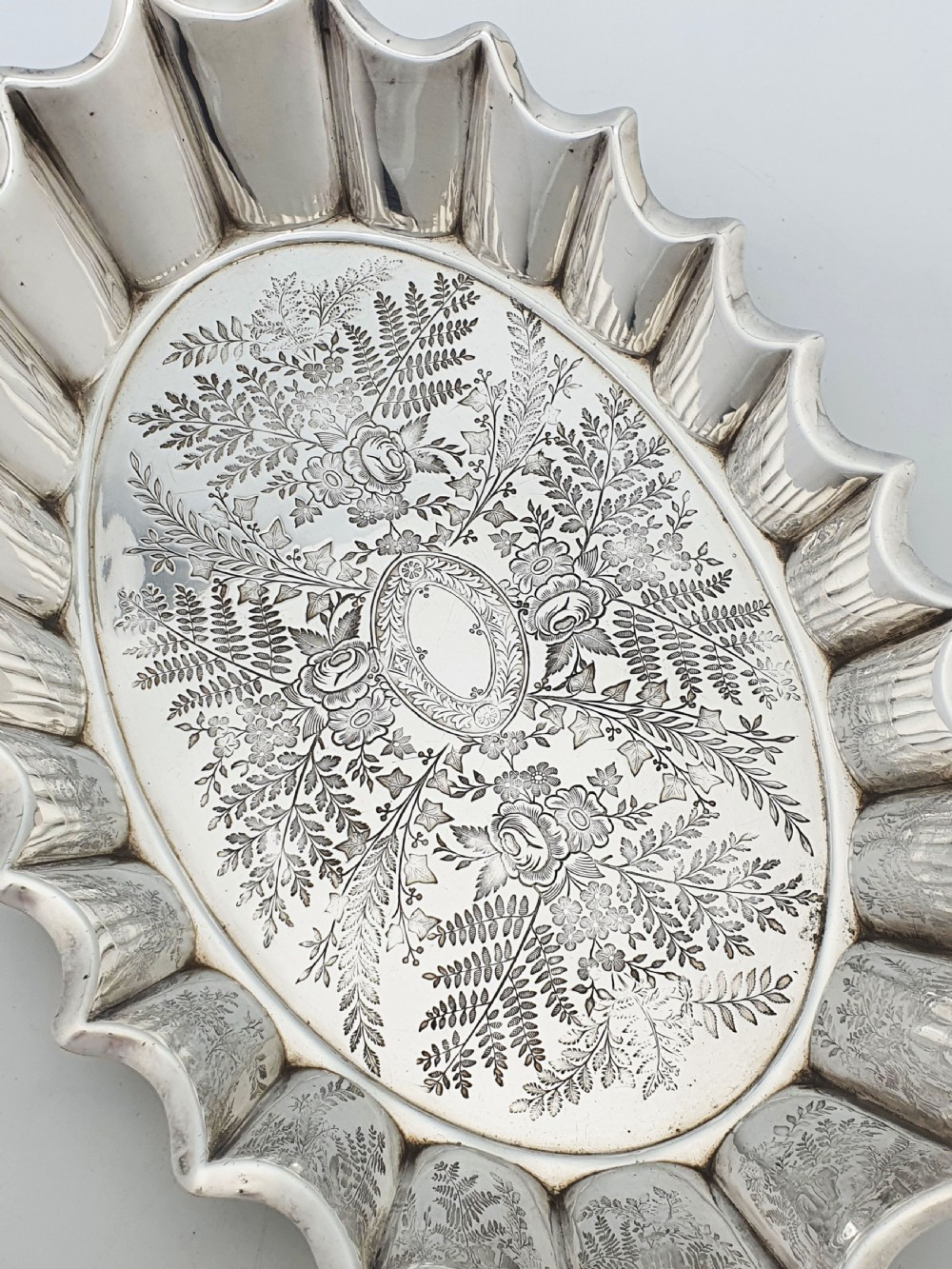 An oval silver plate serving dish featuring intricate floral engravings in the centre and a fluted scalloped outer rim. View II