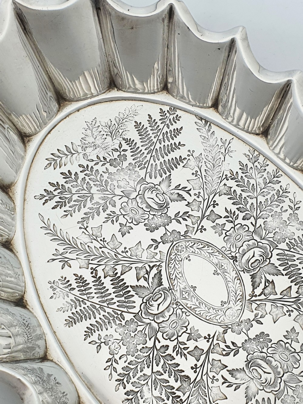 An oval silver plate serving dish featuring intricate floral engravings in the centre and a fluted scalloped outer rim. View III