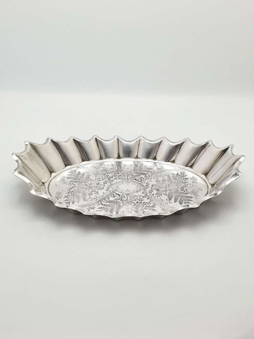An oval silver plate serving dish featuring intricate floral engravings in the centre and a fluted scalloped outer rim. View VII