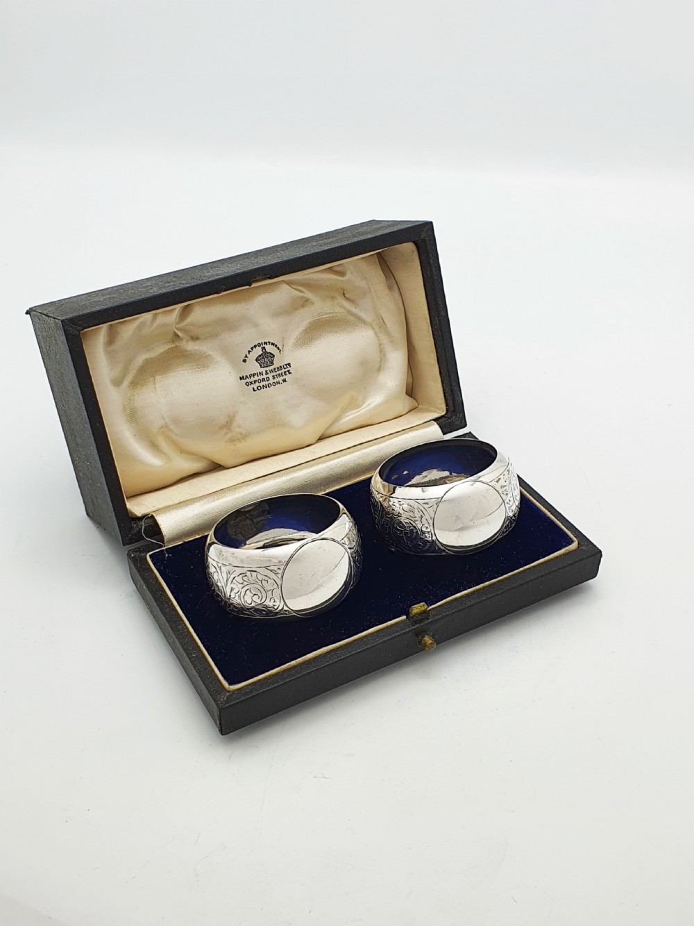 A pair of engraved sterling silver napkin rings with circular cartouches resting inside a dark blue velvet-lined presentation box.