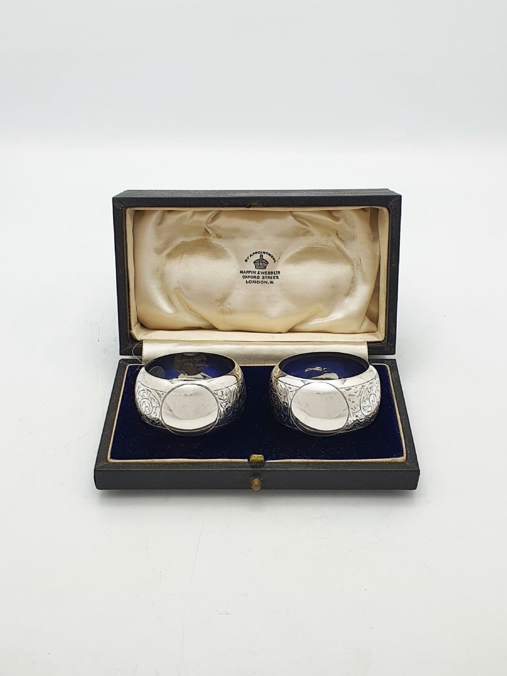 A pair of engraved sterling silver napkin rings with circular cartouches resting inside a dark blue velvet-lined presentation box. View II