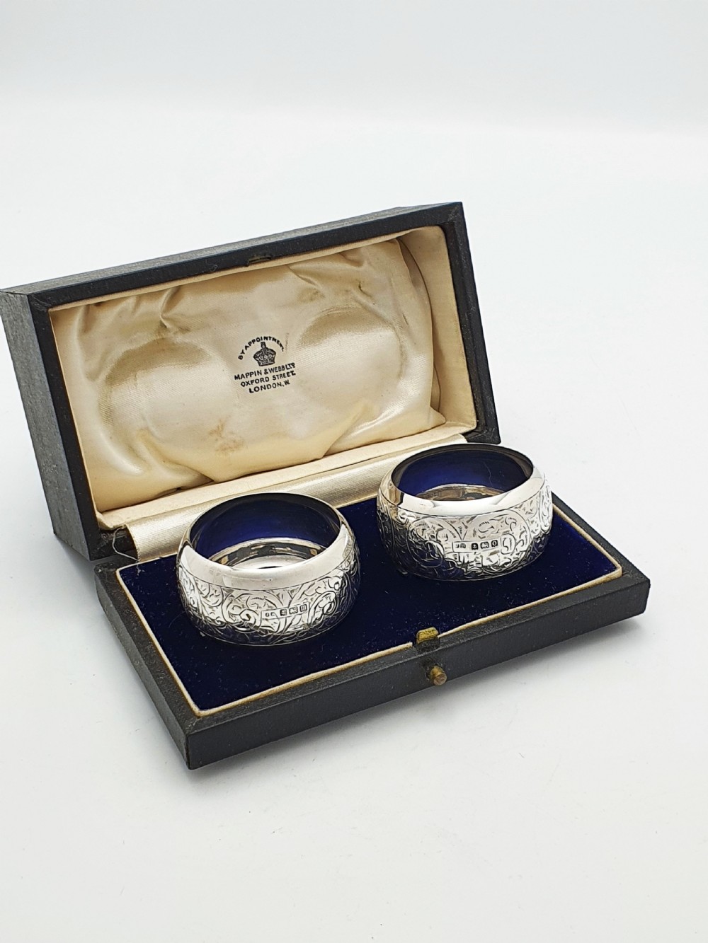A pair of engraved sterling silver napkin rings with circular cartouches resting inside a dark blue velvet-lined presentation box. View III