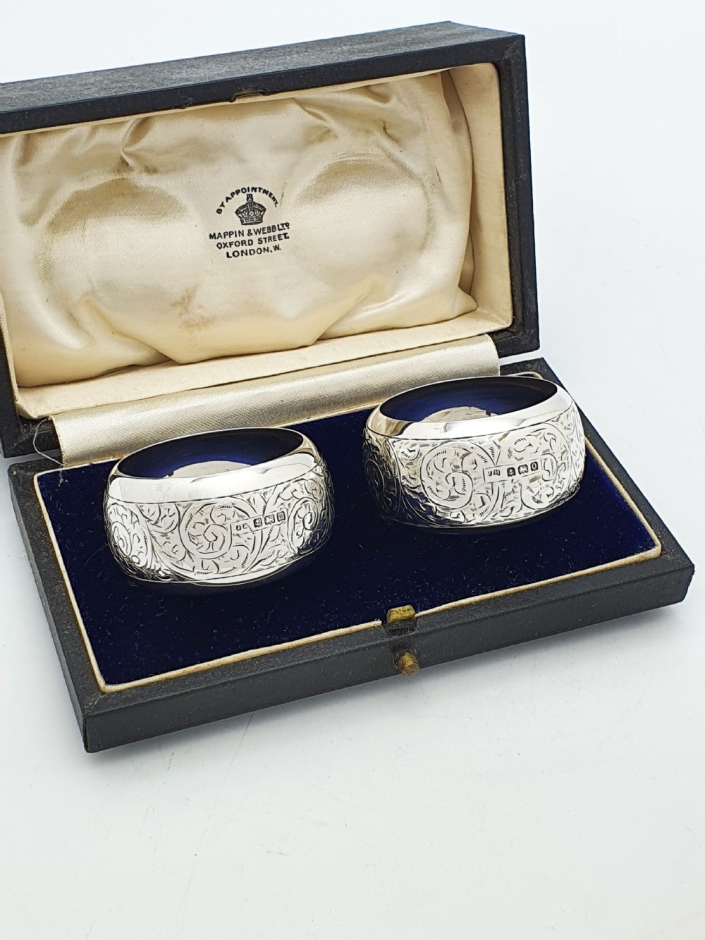 A pair of engraved sterling silver napkin rings with circular cartouches resting inside a dark blue velvet-lined presentation box. View VI