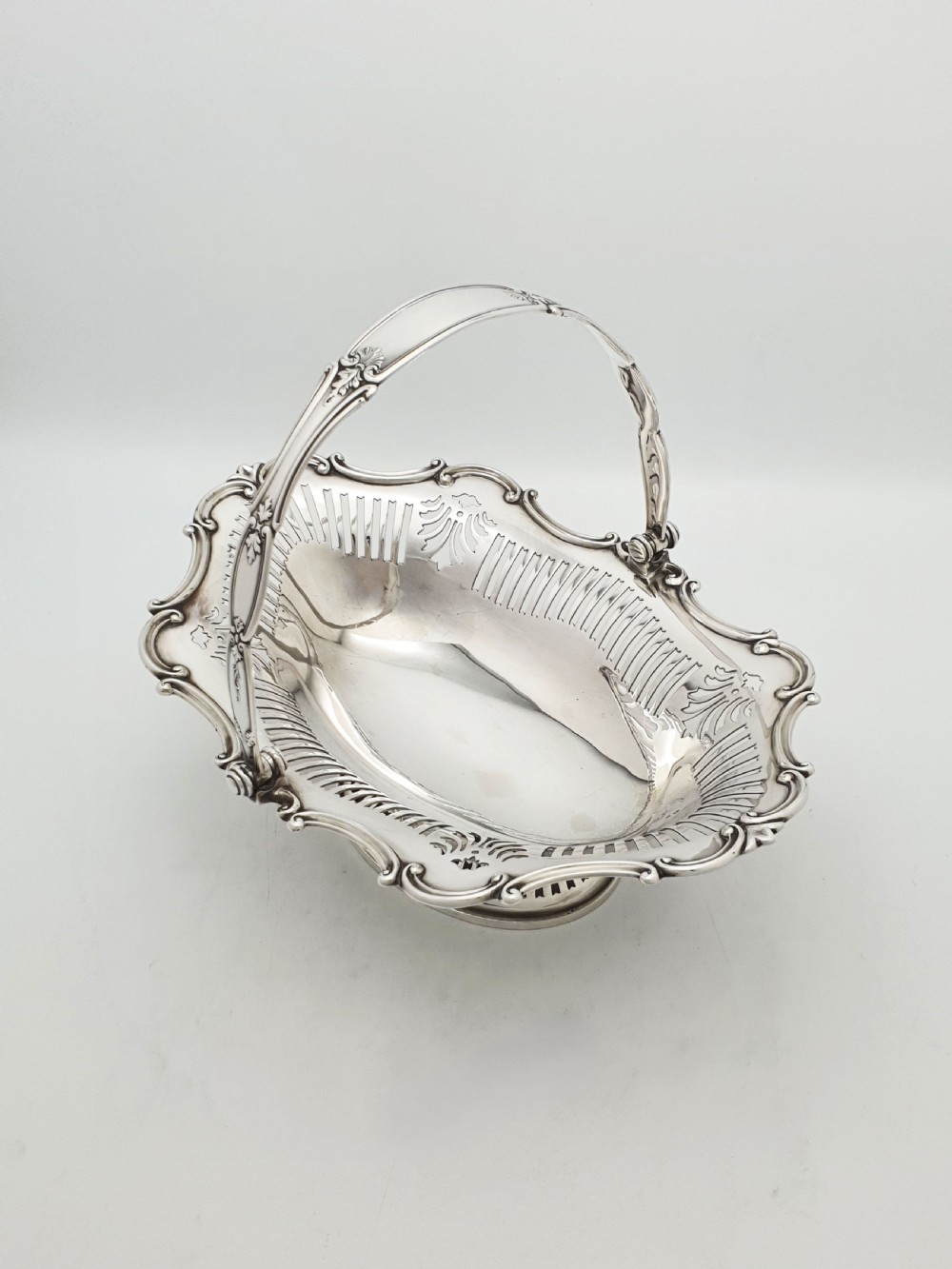 A George V silver-plated oval basket featuring a pierced border, relief rococo scroll edging, and a central hinged swing handle.