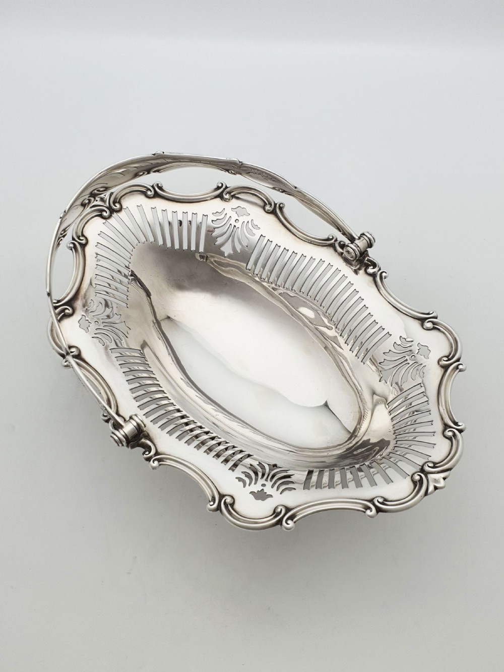 A George V silver-plated oval basket featuring a pierced border, relief rococo scroll edging, and a central hinged swing handle. View V