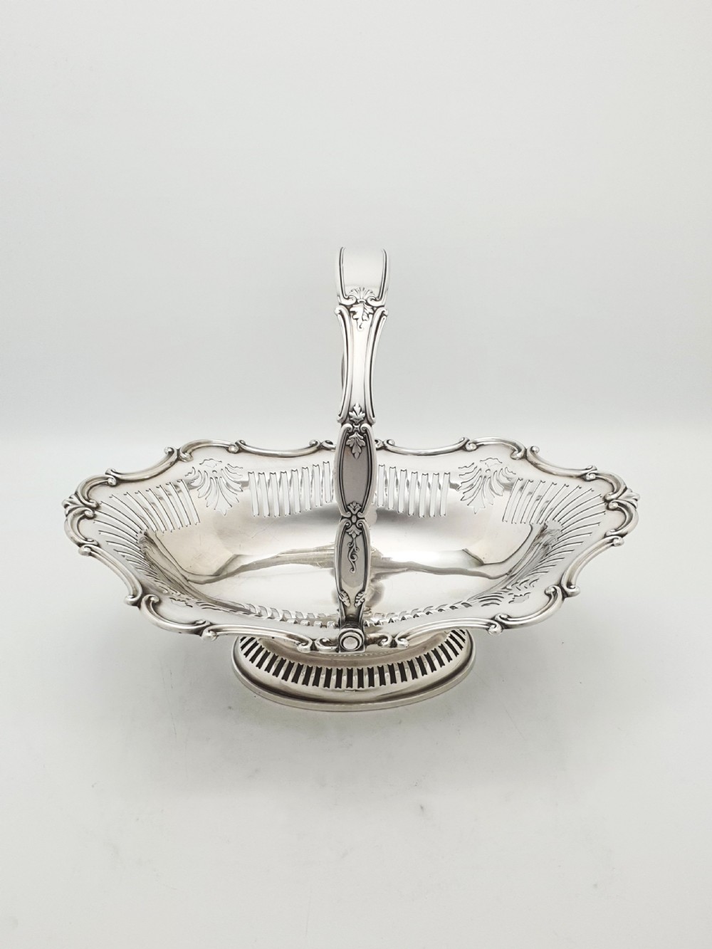 A George V silver-plated oval basket featuring a pierced border, relief rococo scroll edging, and a central hinged swing handle. View VI