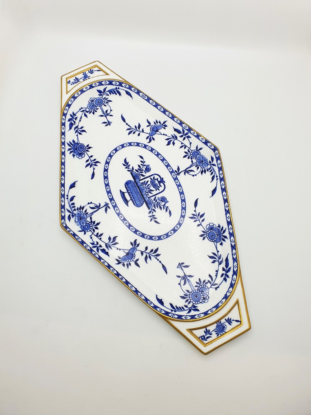 An elongated diamond-shaped white porcelain tray decorated with blue floral transfers, a central floral basket motif, and gilded handles.