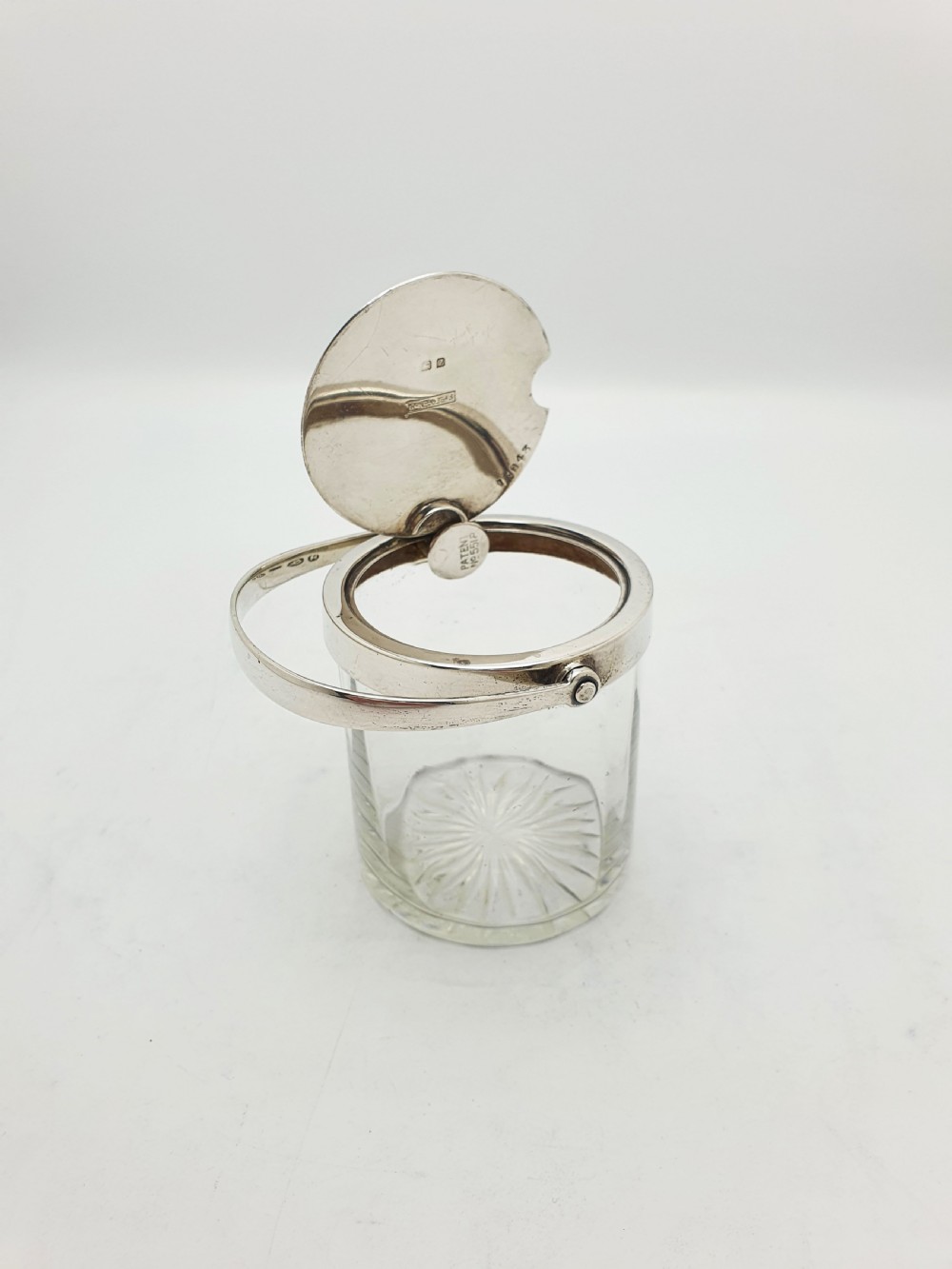 An antique cut glass condiment jar featuring a hallmarked sterling silver rim, swing handle, and hinged lid.