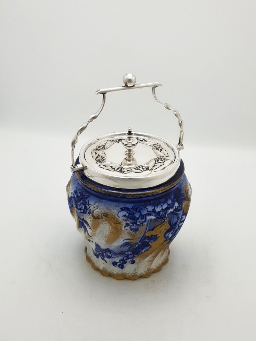 An Edwardian flow blue ceramic biscuit barrel featuring cobalt blue floral motifs, gilded accents, and a silver-plated lid with a twisted swing handle.