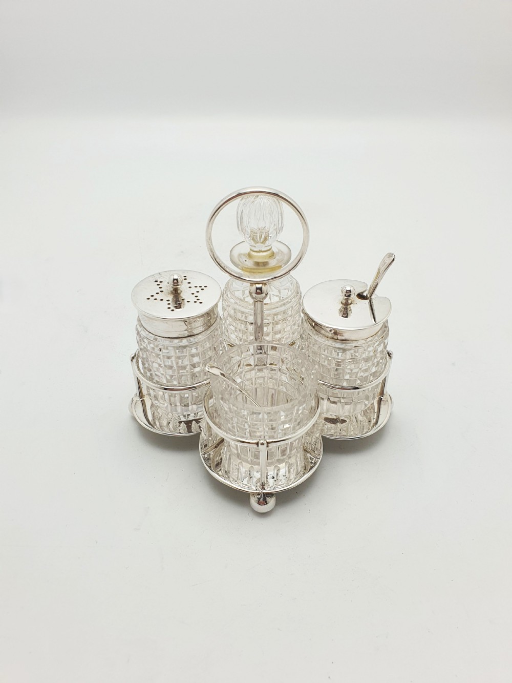 An antique silver-plated condiment caddy holding four faceted cut-glass bottles and pots with silver lids.
