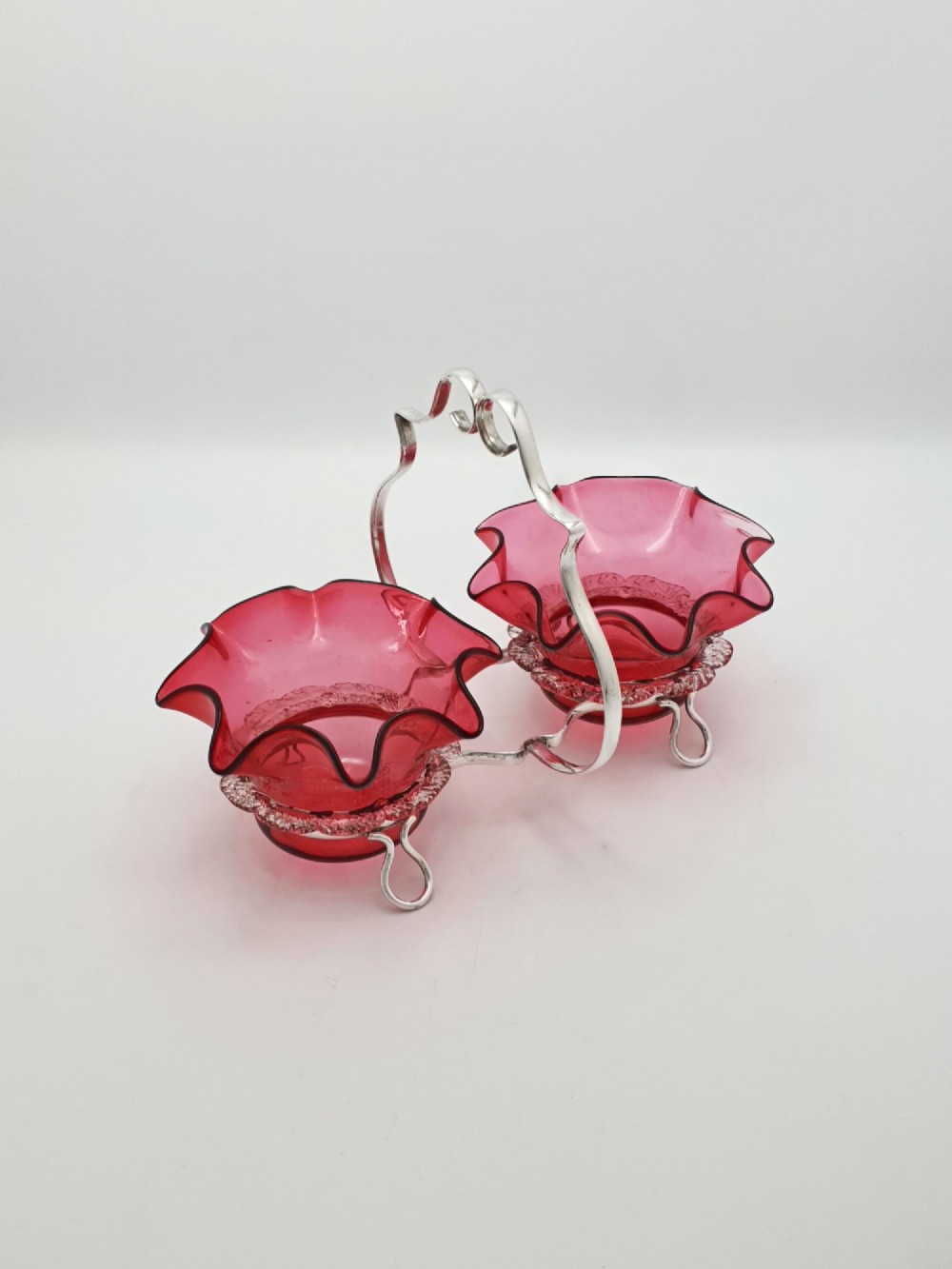 An Edwardian twin cranberry glass preserve dish set with crimped rims held in a silver-plated wireframe stand with a central handle.