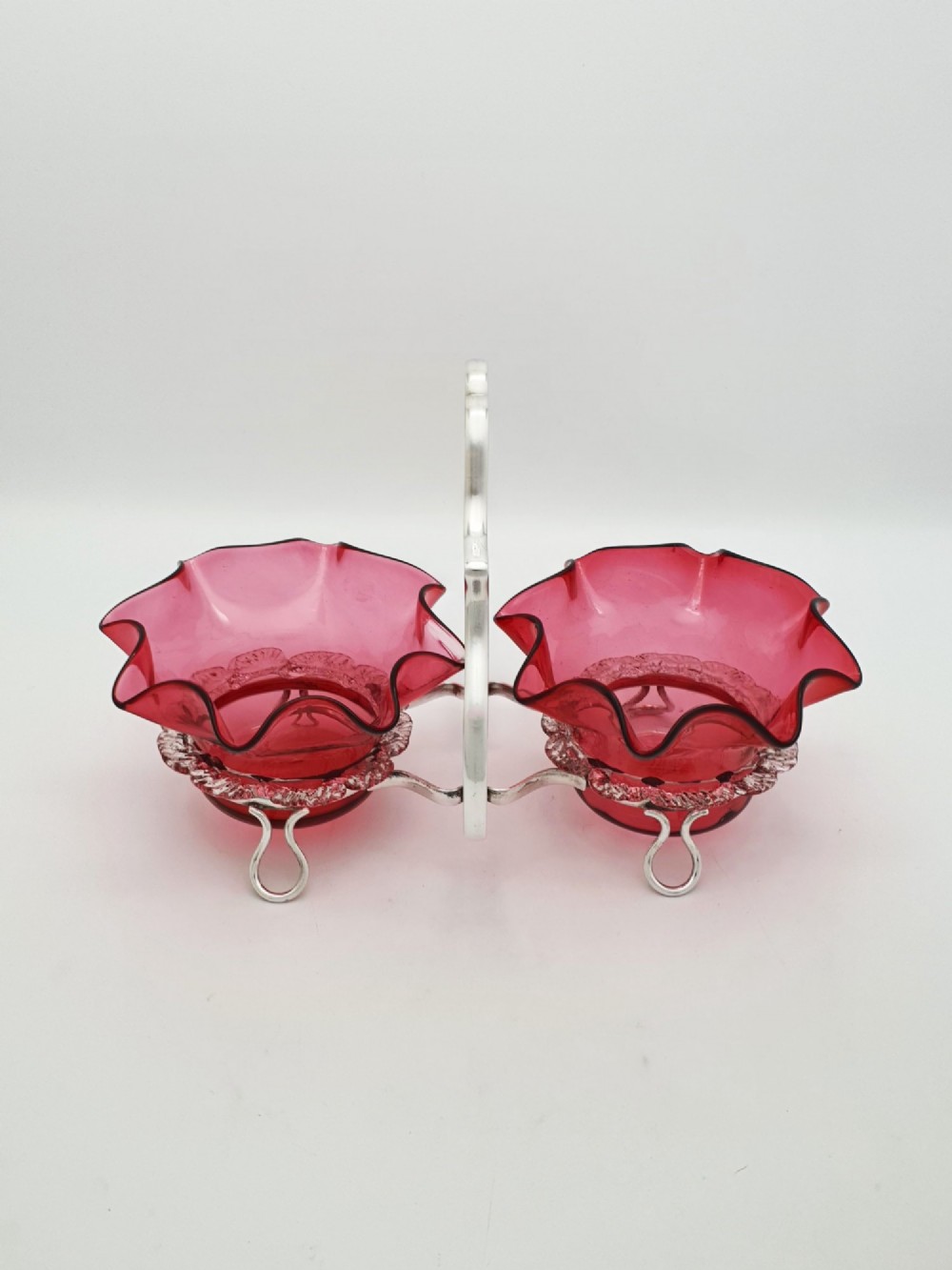 An Edwardian twin cranberry glass preserve dish set with crimped rims held in a silver-plated wireframe stand with a central handle. View VII
