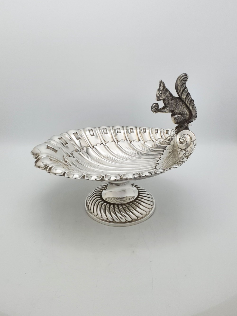 A silver-plated footed shell-shaped dish featuring a figurine of a squirrel perched on the rim holding an acorn, resting on a spiral-lobed pedestal base.
