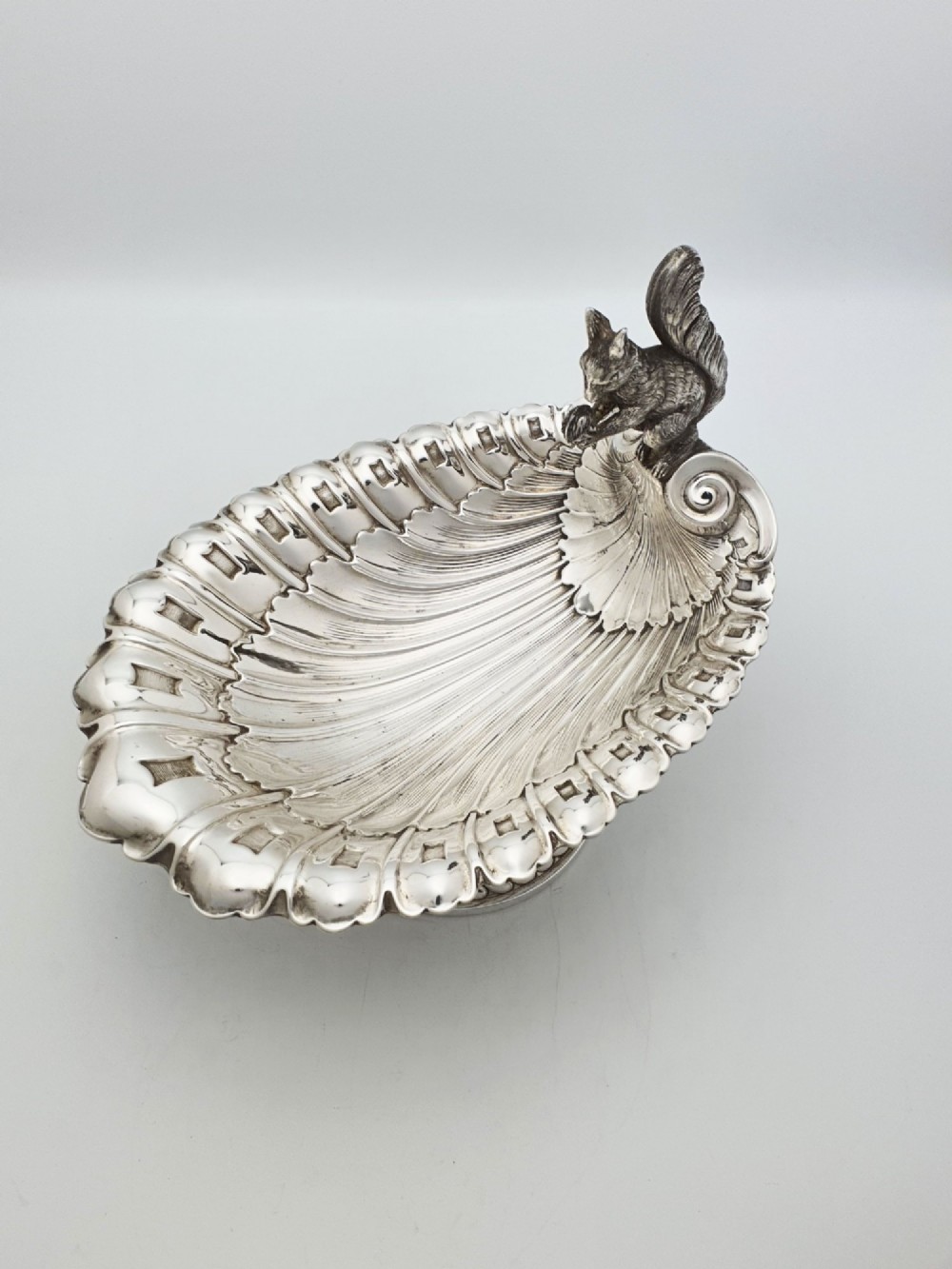 A silver-plated footed shell-shaped dish featuring a figurine of a squirrel perched on the rim holding an acorn, resting on a spiral-lobed pedestal base. View II