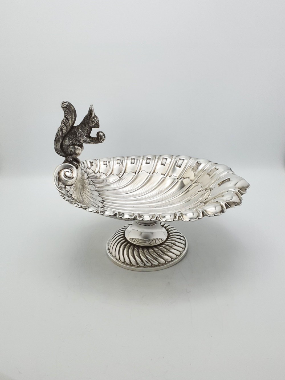 A silver-plated footed shell-shaped dish featuring a figurine of a squirrel perched on the rim holding an acorn, resting on a spiral-lobed pedestal base. View V