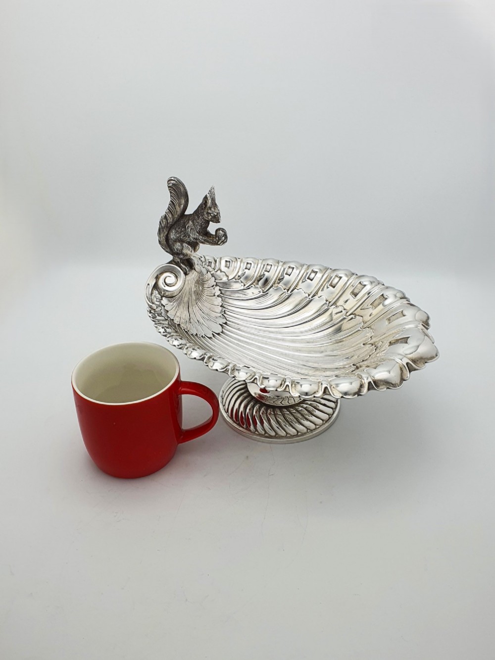 A silver-plated footed shell-shaped dish featuring a figurine of a squirrel perched on the rim holding an acorn, resting on a spiral-lobed pedestal base. View VIII